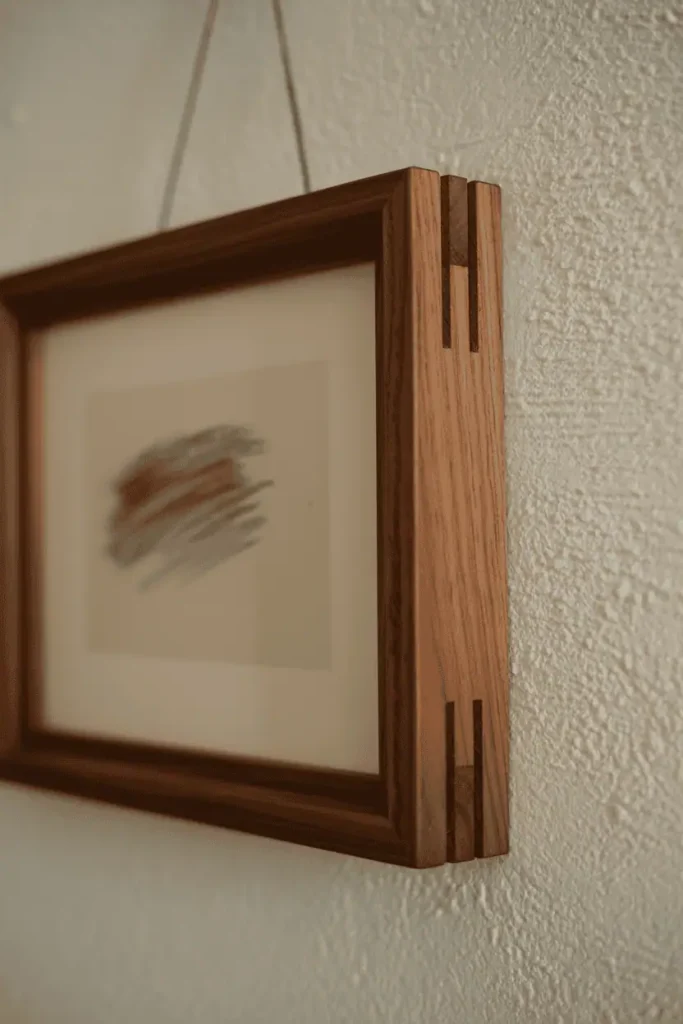 A close-up editorial shot of a single handcrafted wooden frame in walnut with visible dovetail corner joinery. The craftsmanship is clearly shown at the frame corners. Inside: a simple charcoal sketch on cream paper. The frame hangs on a textured plaster wall painted in warm white. Directional light from the side highlights the wood grain and joinery detail. Artisanal, intentional, craft-focused composition.