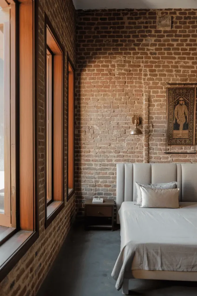 Industrial-heritage Indian bedroom with exposed old brick walls, all window frames and trim in teak wood with minimal carving along edges. Contemporary bed with linen upholstered headboard. Brass sconces, vintage block-printed textile as wall art. Converted haveli aesthetic, urban character. Natural light through teak-framed windows.