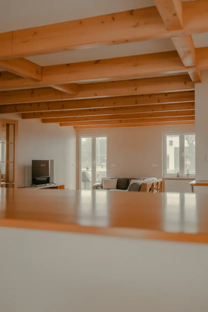 Living room with exposed wooden ceiling beams, neutral walls, minimal furniture, warm natural wood contrast, airy and architectural feel, wide-angle interior view, soft natural daylight, Scandinavian–modern wood aesthetic, realistic interior photography, high resolution, magazine-style composition, shallow depth of field, calm warm palette, no people, no text, no watermark