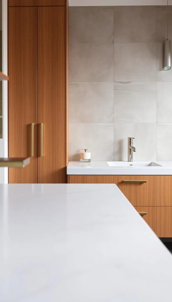 A high-quality contemporary bathroom vanity showcasing a range of modern materials and finishes. In the foreground, a sleek, minimalist vanity countertop in a smooth, lustrous quartz material, reflecting the soft, even lighting. In the middle ground, the vanity cabinet is crafted from warm, natural wood with a matte lacquer finish, complemented by metallic hardware in a brushed nickel or gold tone. Behind, the wall features a striking, textured tile backsplash in a neutral color palette, creating depth and visual interest. The overall scene conveys a sense of sophisticated, spa-like elegance, perfectly suited for a modern bathroom setting.