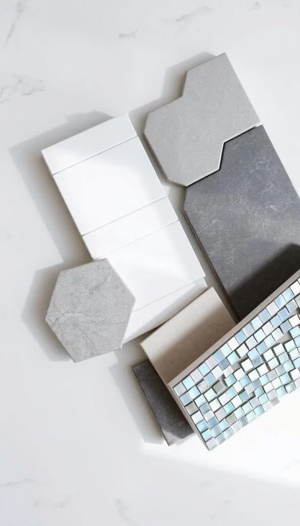 A well-lit, high-angle view of a variety of bathroom tile materials arranged artfully on a clean, neutral surface. The tiles include glossy subway tiles in classic white, matte gray hexagonal tiles, natural stone tiles with subtle veining, and iridescent mosaic tiles in shimmering blues and greens. The tiles are positioned to showcase their unique textures, patterns, and colors, creating an inviting and aspirational display. The lighting is soft and diffused, highlighting the depth and nuance of each material. The overall mood is one of serene sophistication, hinting at the transformative potential of these tiles to elevate a bathroom space.